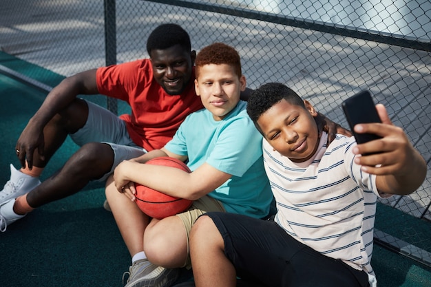 Content black boy sitting on ground and using smartphone while taking selfie with father and brother after basketball