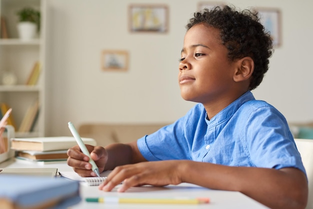 Content Black boy with curly hair sitting at table and making notes in workbook while watching online lesson