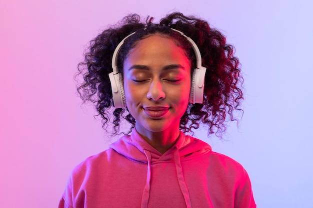 Content black lady enjoying music with in headphones pink hoodie and closed eyes