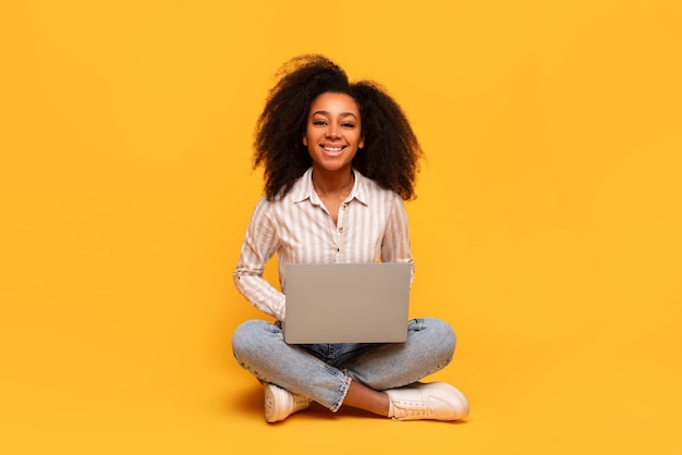 Content black woman with laptop sitting crosslegged on yellow backdrop