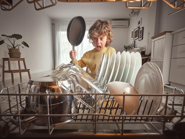Довольный мальчик со сковородой в руках, стоящий возле открытой посудомоечной машины с различной чистой посудой во время домашних дел на кухне