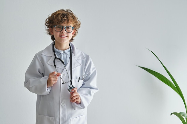 Content child in eyewear and medical robe with professional instrument looking at camera on white background