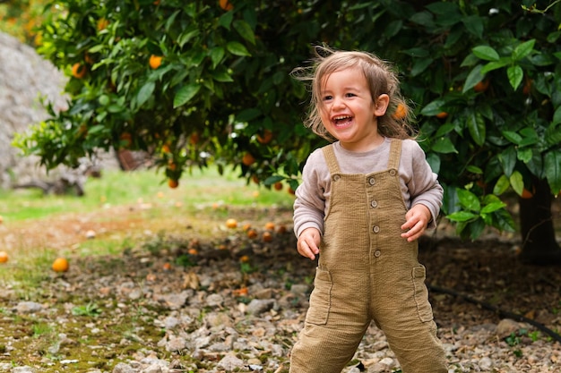 Content child in overalls laughing on pebble terrain and looking away against lush tree with oranges in countryside