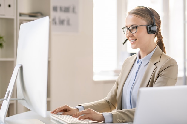 Content confident attractive young call center operator in headset with microphone sitting at table and typing on computer while answering call