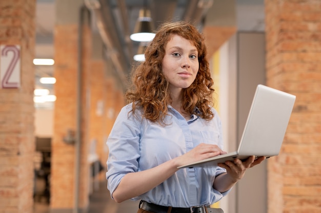 Content confident young woman with curly red hair standing in loft co-working space and using laptop