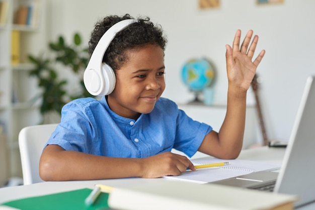 Contenuto carino ragazzo afroamericano in camicia blu seduto al tavolo e agitando la mano allo schermo del laptop whi