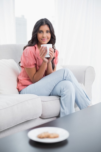 Content cute brunette sitting on couch holding cup of coffee