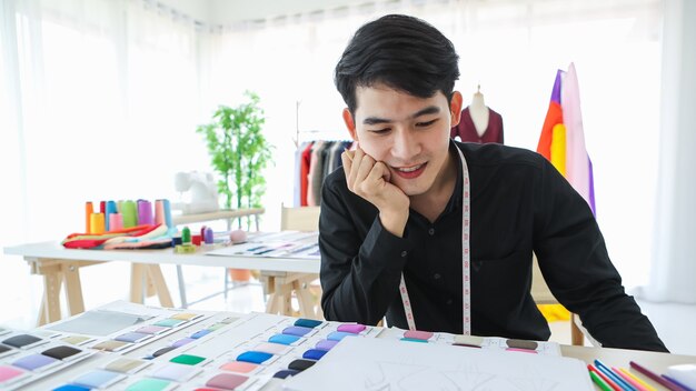Дизайнер содержимого этнической мужской одежды, сидящий за столом с тканью и эскизами с счастливым лицом.