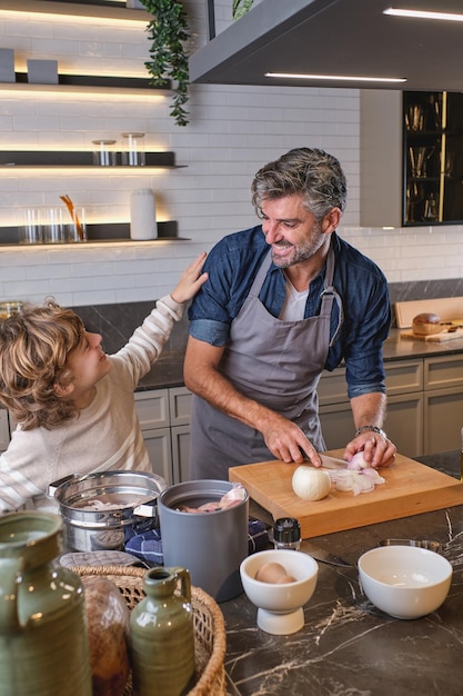 Content father cutting fresh onion near kid while looking at each other at table in light kitchen during cooking process