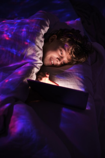 Content jongen surfen tablet met een glimlach terwijl liggend in bed onder de deken in de slaapkamer met zwak licht 's nachts
