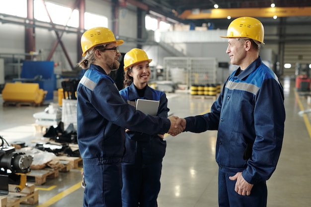 Content mature factory executive in hardhat congratulating workers with workplan implementation and shaking hand of one of them in warehouse