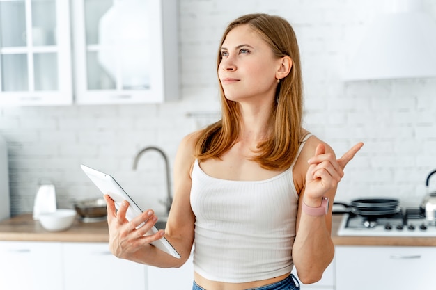 Content peaceful beautiful woman using digital tablet standing in the kitchen at home. Counting with fingers, reading receipes. Healthylife concept.