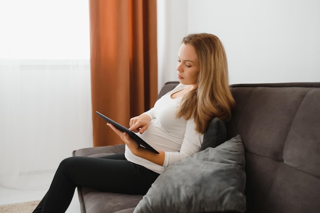 Content pregnant woman holding her tablet sitting on a couch in the living room at home