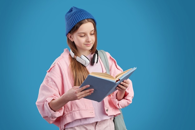 Content schoolgirl with headphones around neck standing and reading textbook