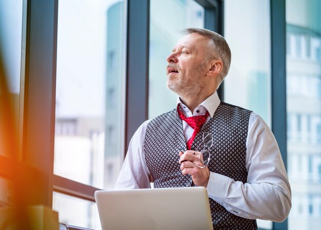 Content senior business man sitting in a suit Laptop in hands Red tie Confident Man looking at the window Business style concept