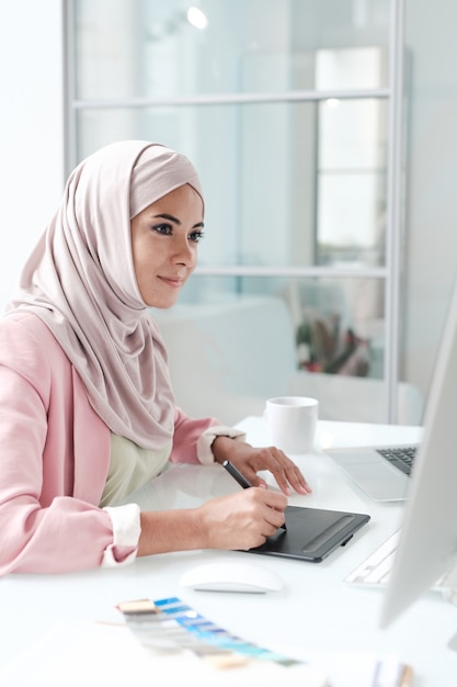 Content young Arabian designer in hijab sitting at table and making pictures on drawing pad