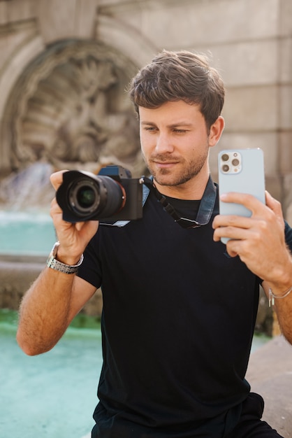 Content young guy using smartphone and photographing on camera near fountain