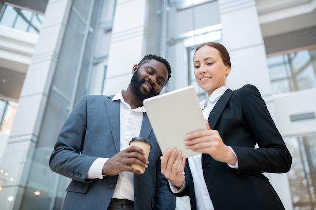 Content young multi-ethnic colleagues in suits standing in office and using tablet while discussing online report