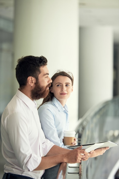 バルコニーに立って同僚の話を聞いているフォルダーとテイクアウトのコーヒーを持つコンテンツの若い女性