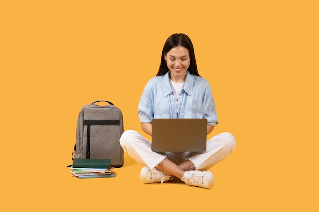 Удовлетворенная студентка с ноутбуком и рюкзаком на ярко-желтом фоне
