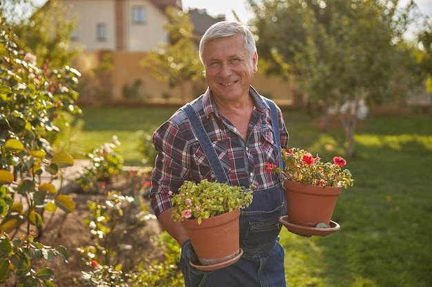 Giardiniere contento che sorride mentre tiene in mano vasi di fiori e sta fuori in giardino