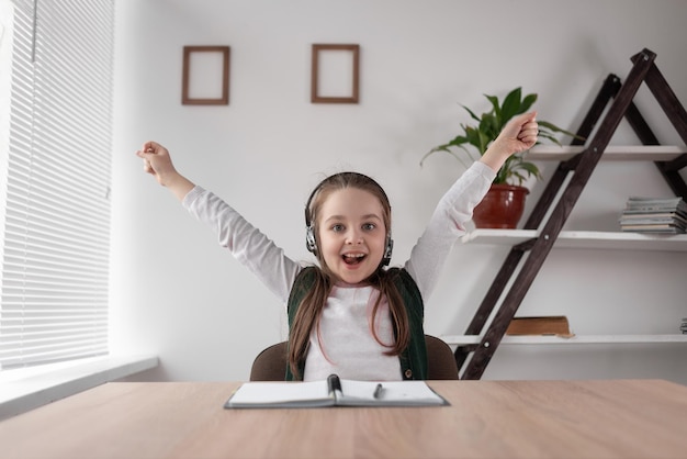 A contented happy child sitting at home at a desk in wireless headphones rejoices at the end of a video tutorial with a teacher Distance learning and loneliness Good news hurray vacation