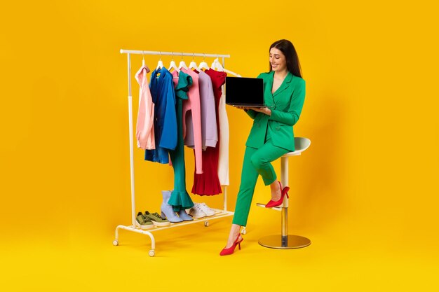衣料品のレールの近くに座って新しいストアのウェブサイトを宣伝する空白の画面でラップトップを示す満足している女性