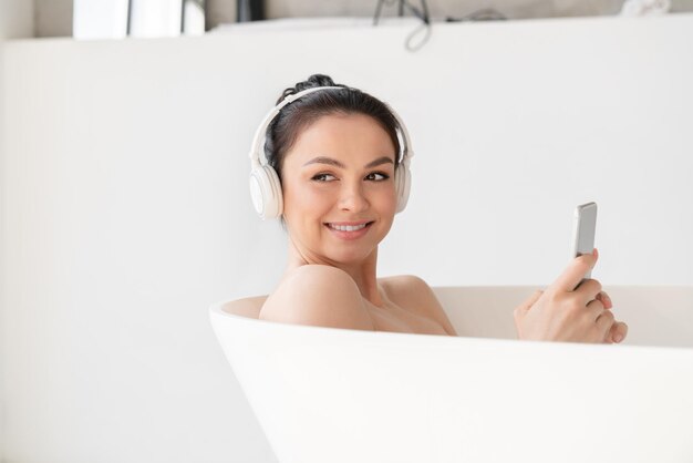 Contented young woman relaxing in hot warm bath shower after hard day listening to the music in headphones using smart phone Beauty treatment skin care Spa procedures concept