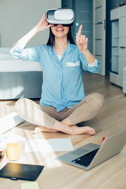 ラップトップを横に置いて床に座って、仮想現実にいる間に見えないものを指している満足している若い女性