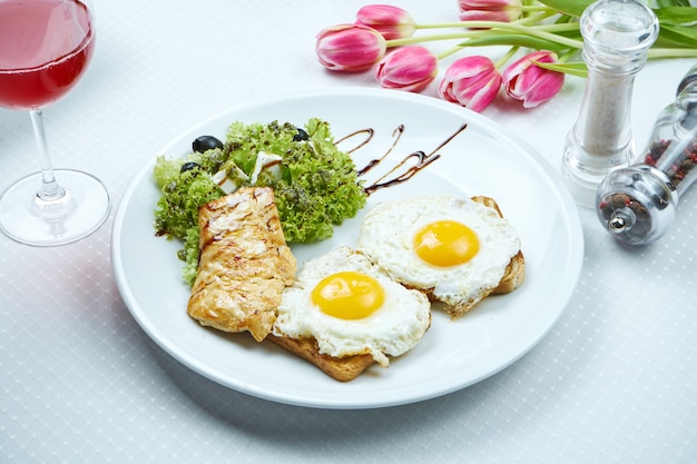 Continental breakfast - toasts with fried eggs, grilled chicken filet, salad with feta and olives on a white plate. Top view, copy space