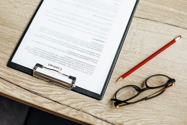 A contract on a wooden work table with glasses and a red pencil