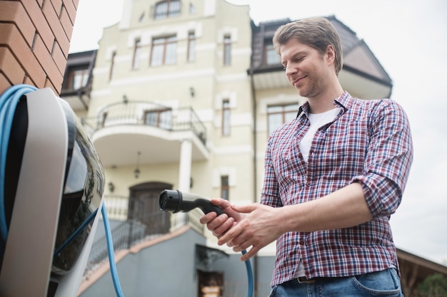 Convenience. Satisfied man in plaid shirt standing outdoors on territory of house near charging station for electric car