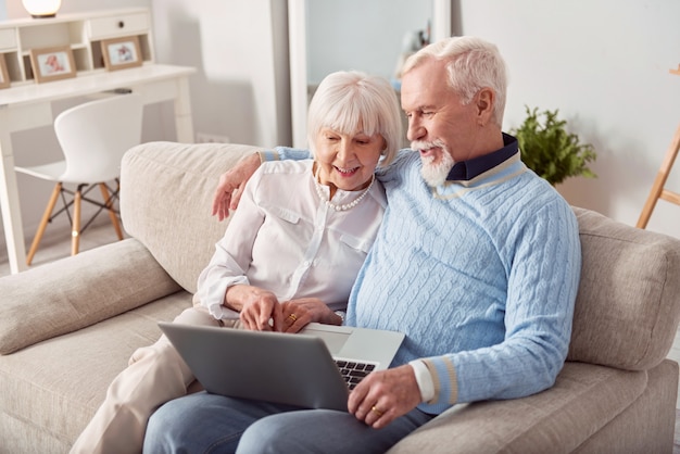 Acquisto conveniente. felice coppia di anziani seduti sul divano in soggiorno e scegliendo un nuovo laptop nel negozio online insieme