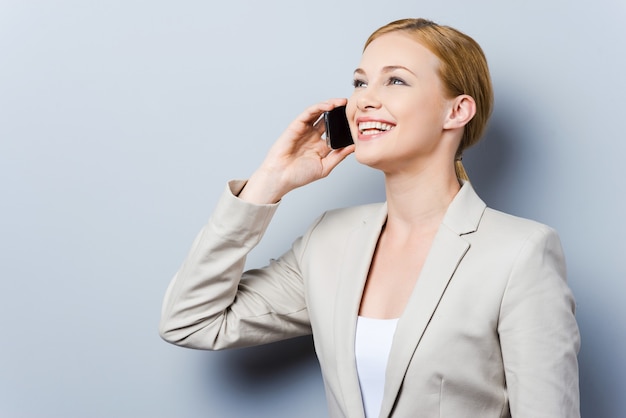 Conversation with pleasure. Beautiful young businesswomen talking on the mobile phone and smiling while standing against grey background