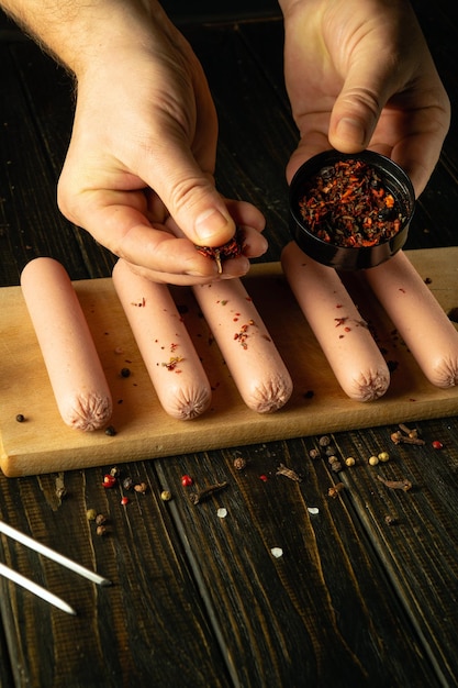 The cook adds spices to raw meat sausages before grilling Concept of cooking fried Munich sausages on kitchen table by chef hands