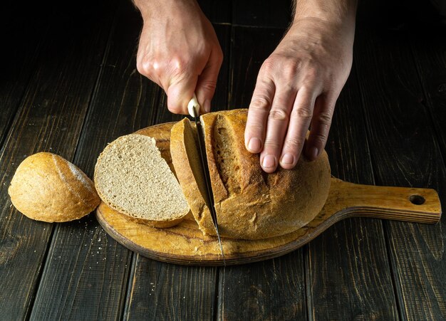 The cook cuts the baked bread with a knife on the cutting board of the restaurant kitchen