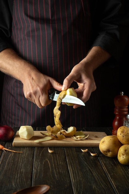 The cook cuts raw potatoes into pieces with a knife before preparing breakfast or dinner