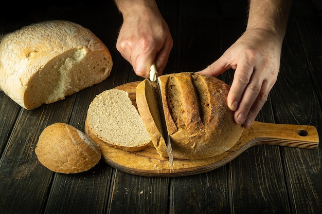 The cook cuts wheat bread with a knife on a cutting wooden board