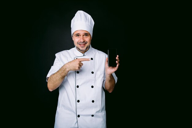 Cook dressed in white hat and jacket pointing at his mobile phone on black background. Restaurant, food and takeaway concept.