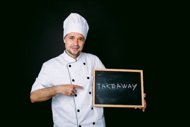 Cook dressed in a white jacket and hat holding a blackboard that reads: "Take away" on a black background. Restaurant and takeaway concept.