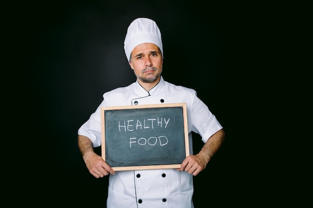 Cook dressed in a white jacket and hat holds a blackboard that reads: "Vegan" on a black background. Healthy food and restaurant concept.