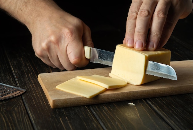 Le mani del cuoco con il coltello tagliano un formaggio sulla tavola di legno per sandwich pizza italiana o spuntino in cucina preparazione per cucinare mangiare sano e stile di vita concetto di cibo