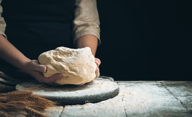 cook holds the dough over the cutting board amid flour and wheat on a black background.