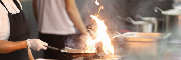 Cook in kitchen holds frying pan in which fire is burning.