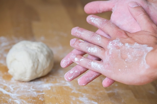Cook shakes off flour from his hands against the background of a lump of dough on a wooden table