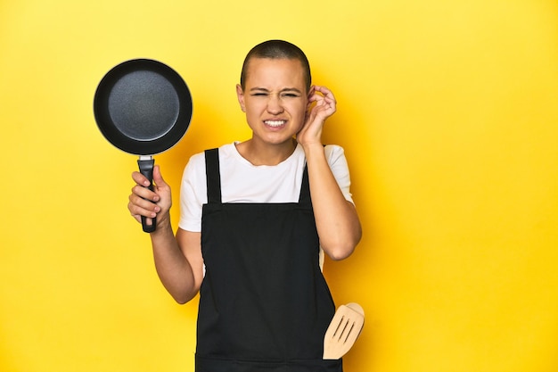 Cook woman holding a pan yellow studio background covering ears with hands