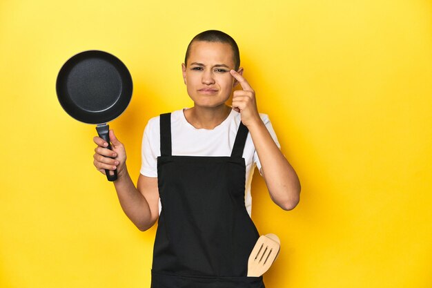 Cook woman holding a pan yellow studio background showing a disappointment gesture with forefinger