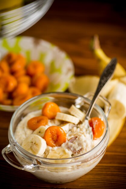 cooked boiled sweet oatmeal with with dried apricots and bananas in a bowl on a wooden table
