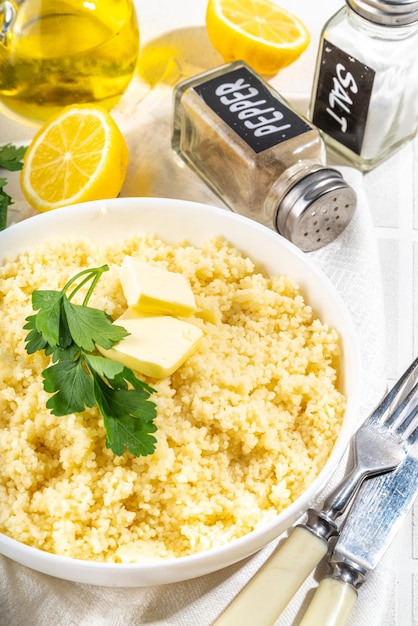 Cooked couscous with cilantro or parsley butter olive oil lemon in white ceramic bowl on kitchen white table copy space