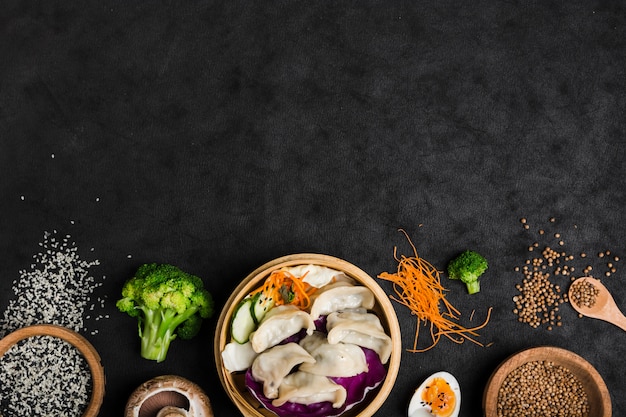 Cooked dumplings inside the bamboo steamer with eggs; broccoli; sesame and coriander seeds on black texture background